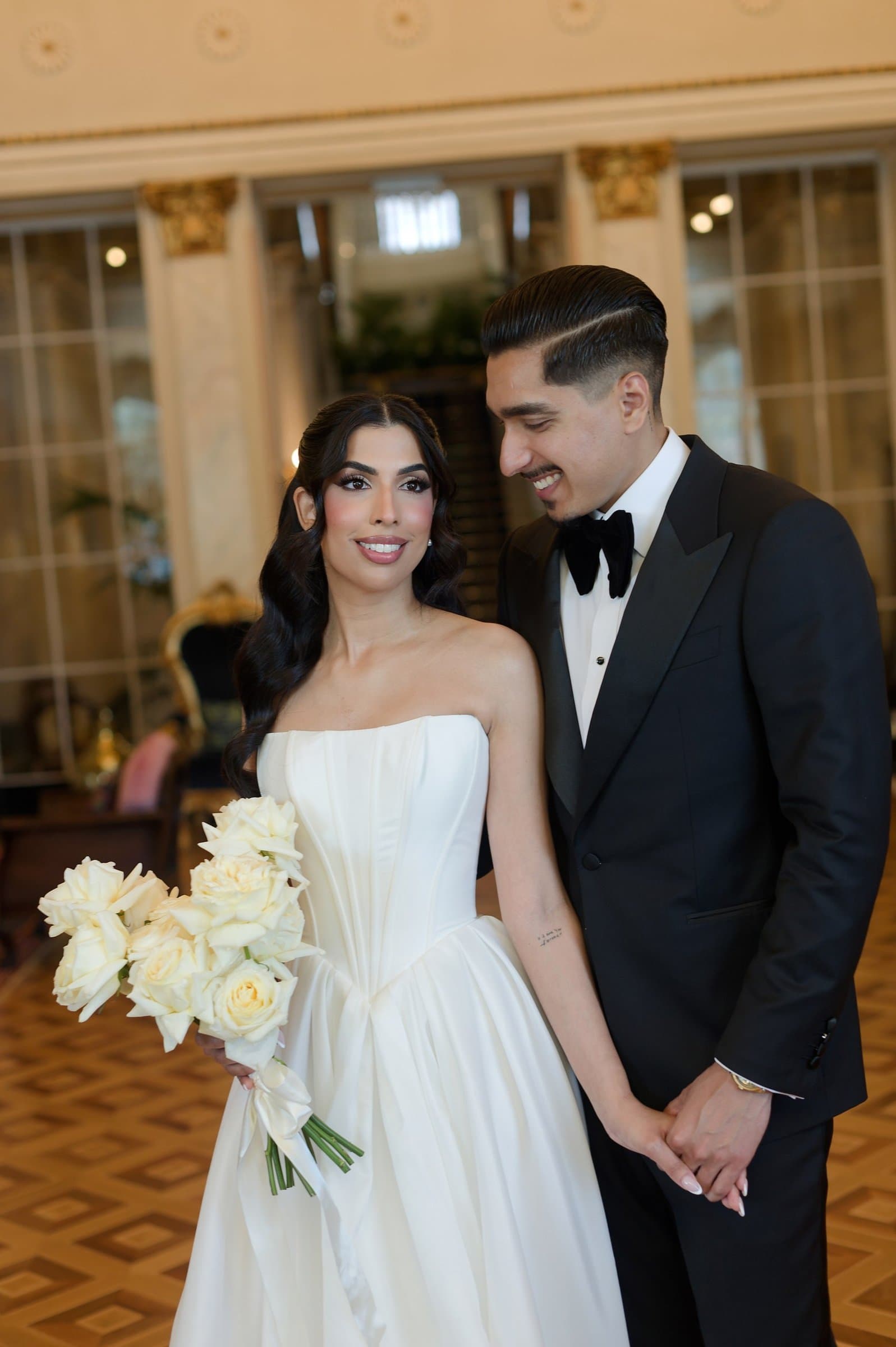 The bride and groom in a hotel hall, she holding white roses, he in a black tuxedo