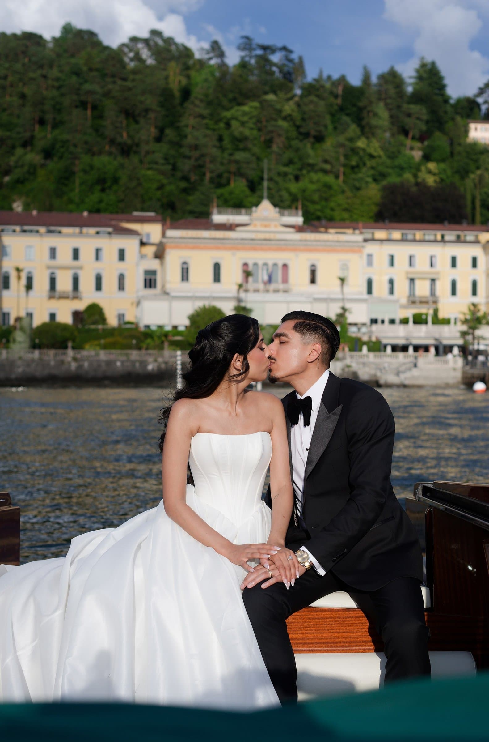 The bride and groom kissing aboard a wooden boat, a lakeside grand hotel behind