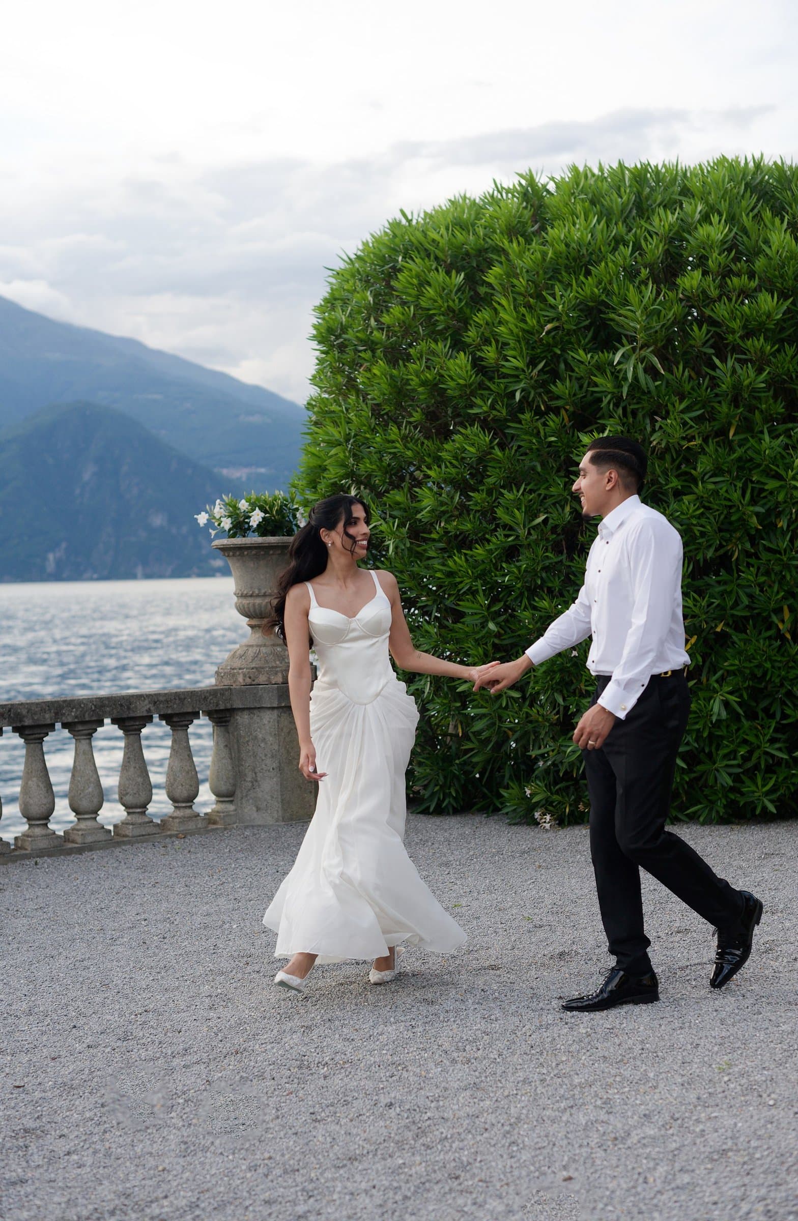 The couple walking hand in hand on a lakeside terrace, the groom in a white shirt and black trousers