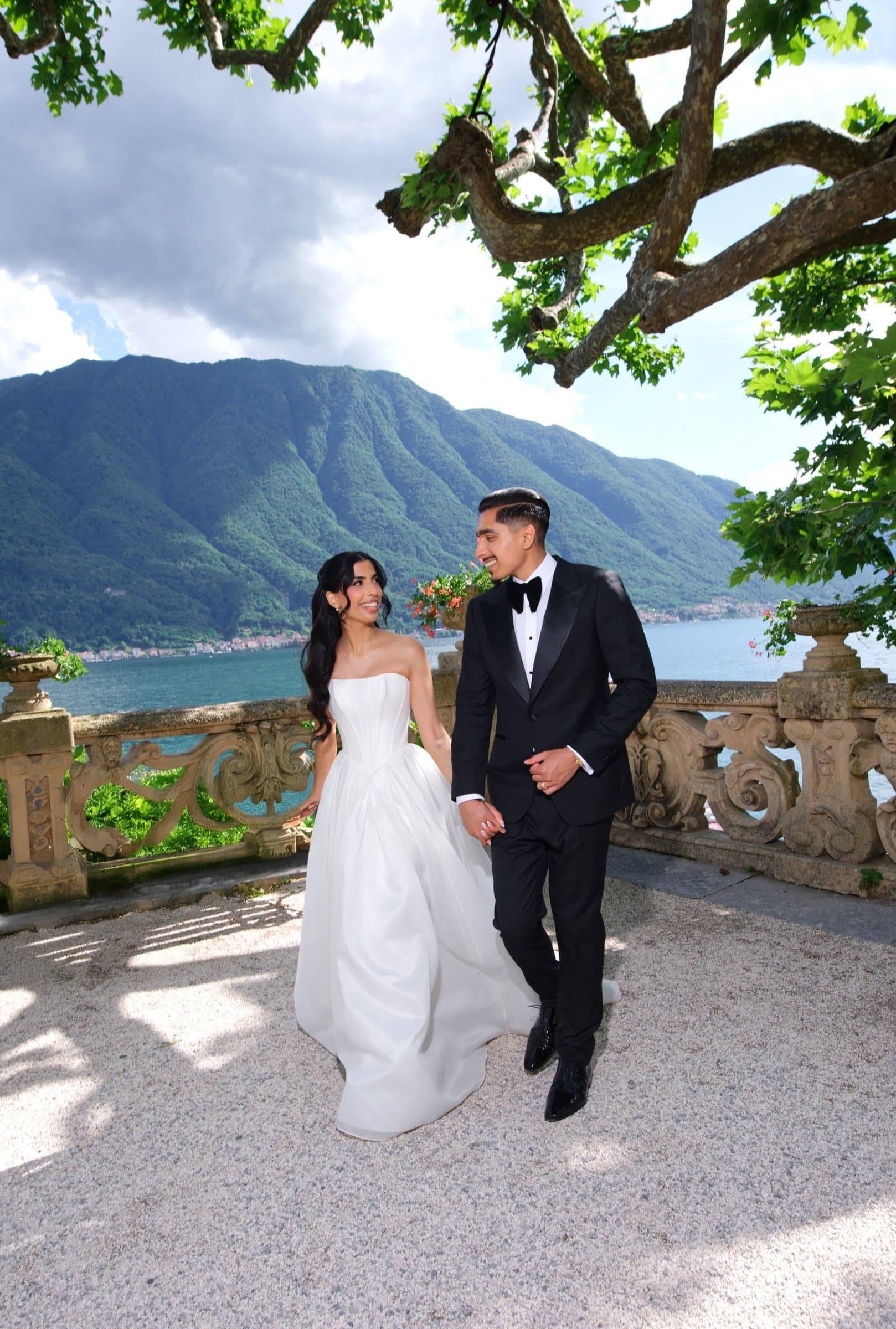 The bride and groom walking along a villa balustrade under a plane tree, Lake Como behind
