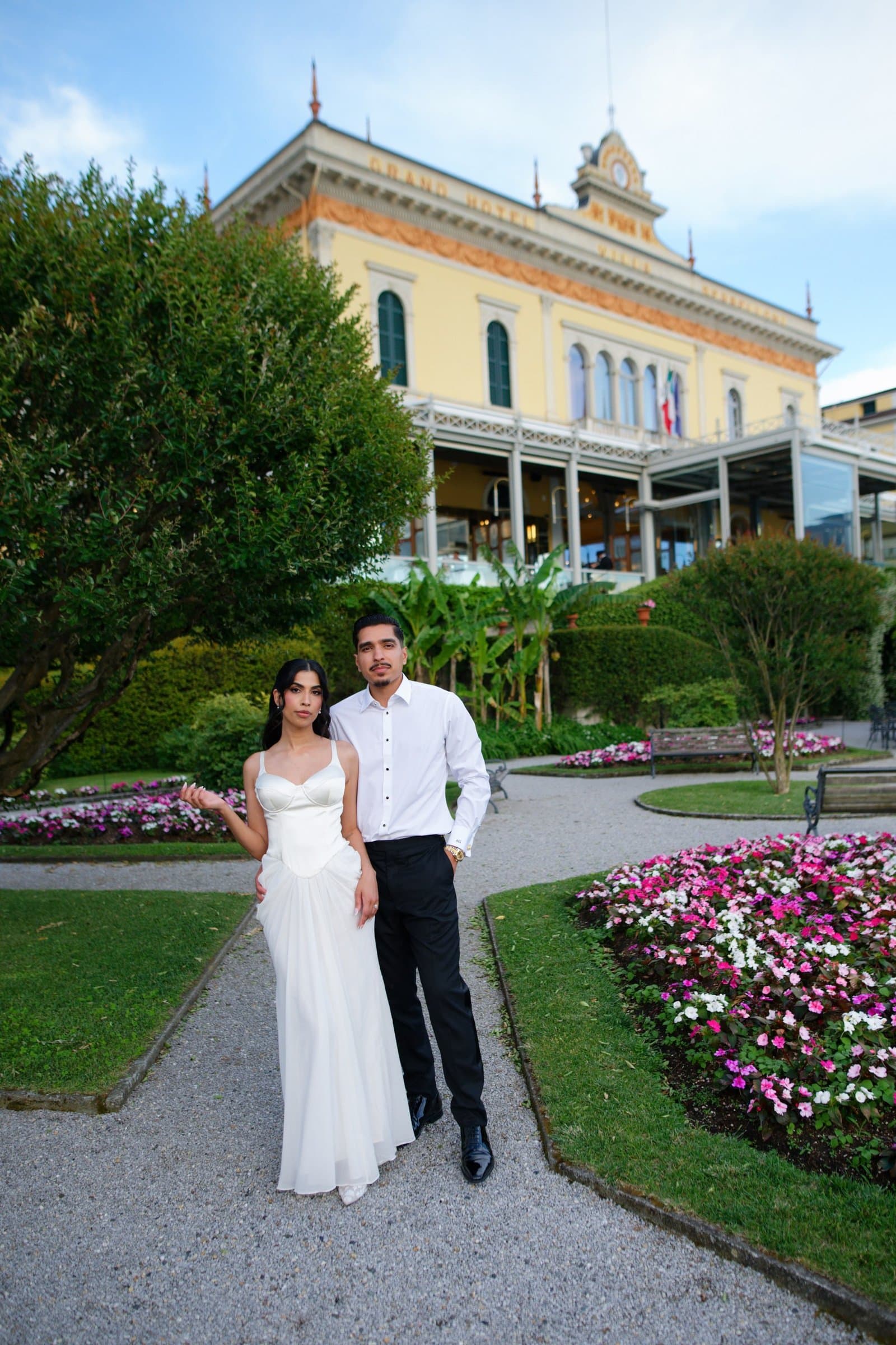 The couple on a garden path in front of a lakeside grand hotel