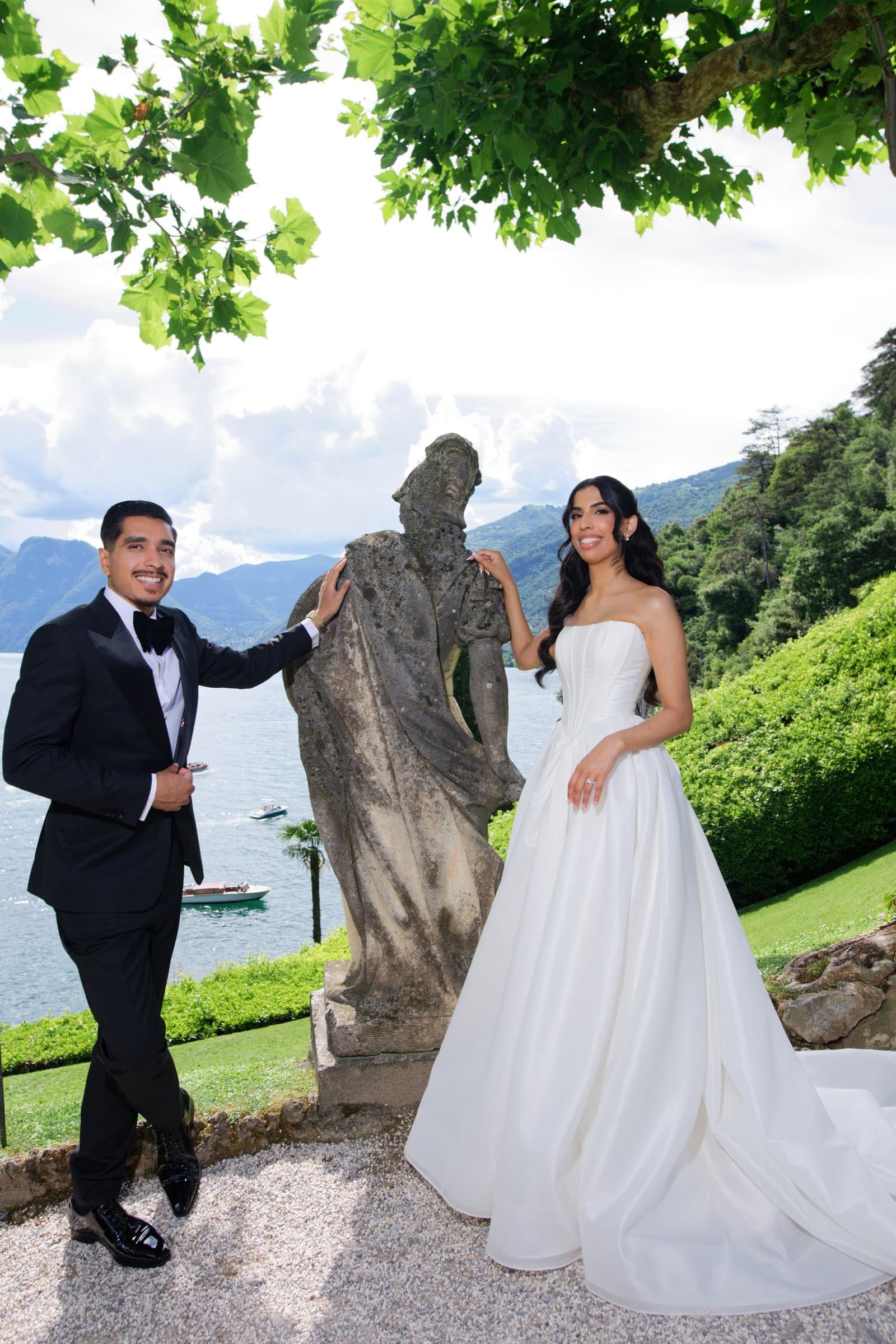 The groom in a black tuxedo beside the bride and a garden statue above Lake Como