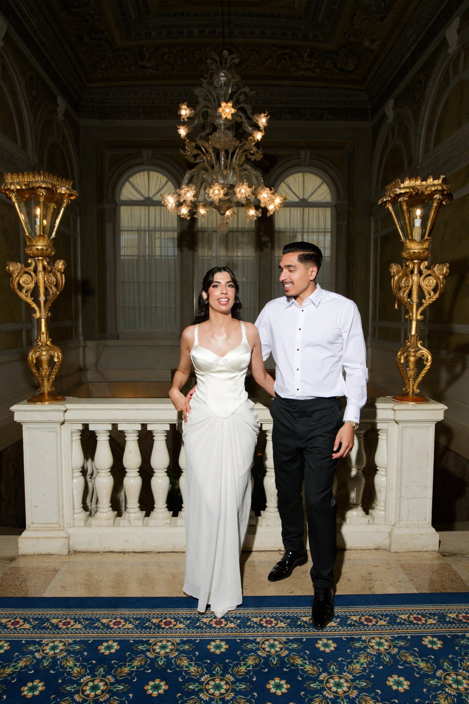 The couple in a gilded hall under a lit chandelier, dressed for the evening