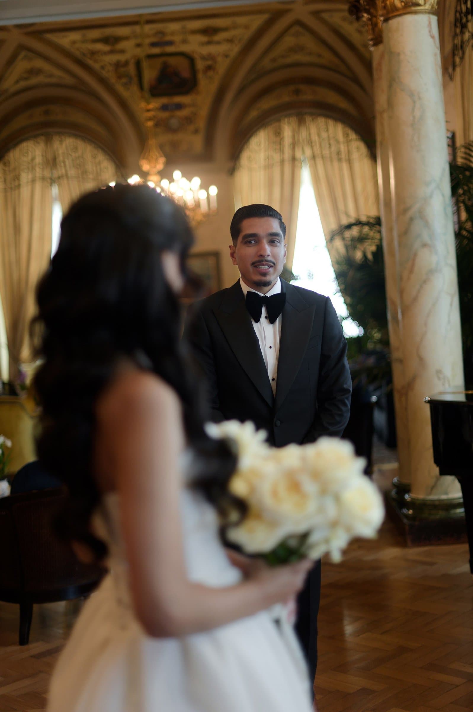 The groom in a black tuxedo in a gilded hotel salon, the bride and her white roses in the foreground