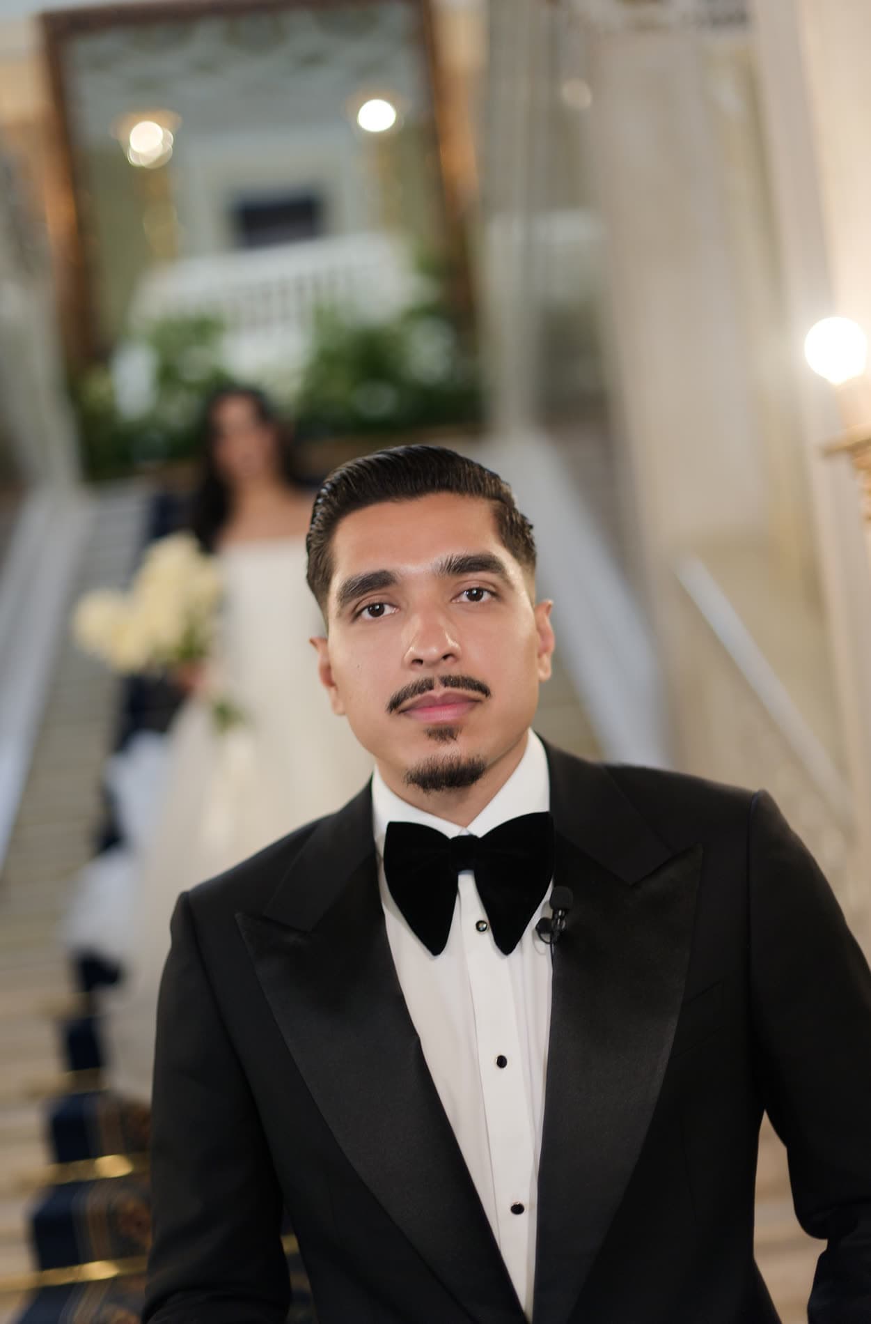 A groom in a black custom tuxedo and bow tie, his bride on the staircase behind him