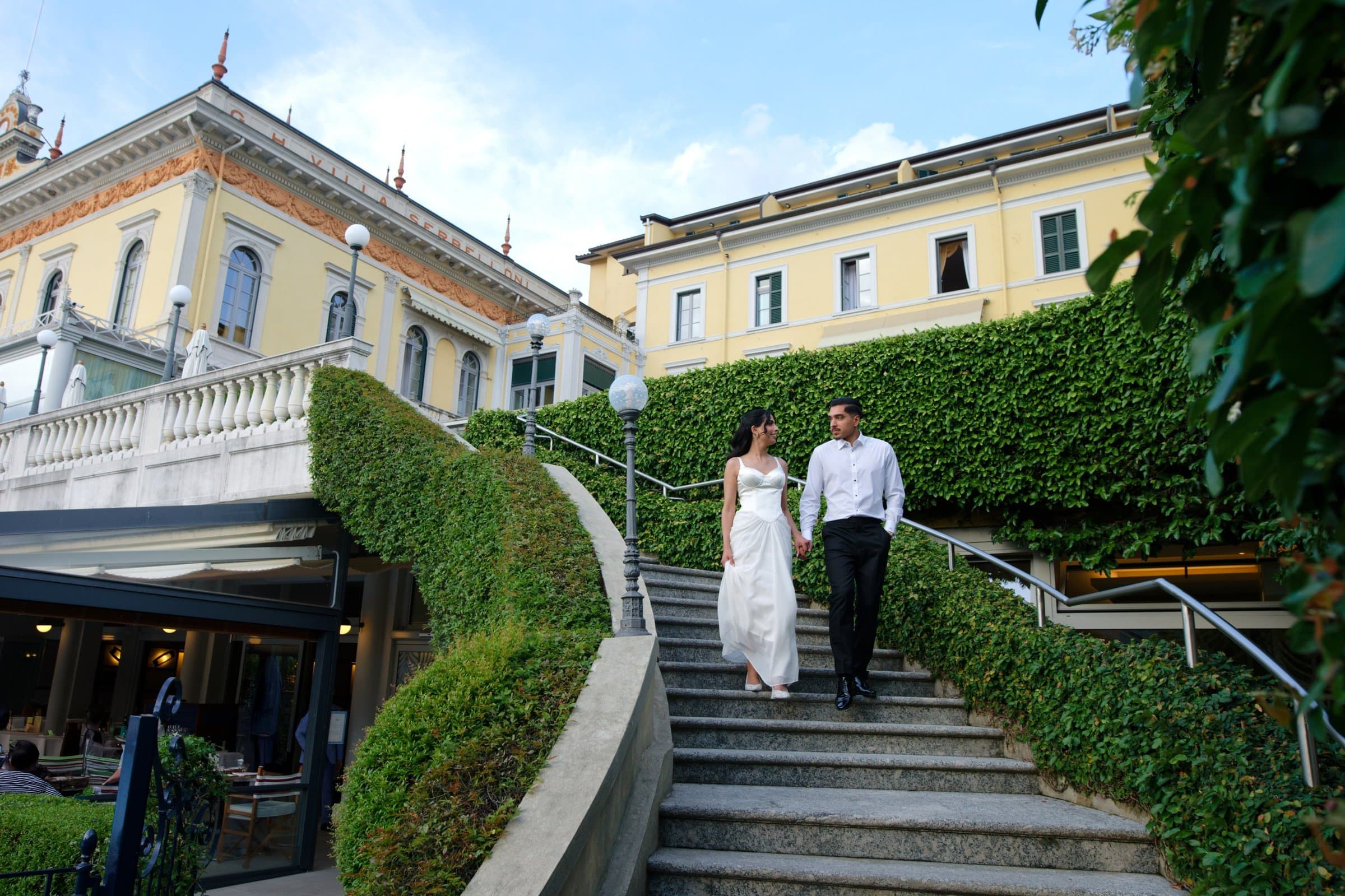 The bride and groom walking down hedge-lined steps from a grand hotel terrace