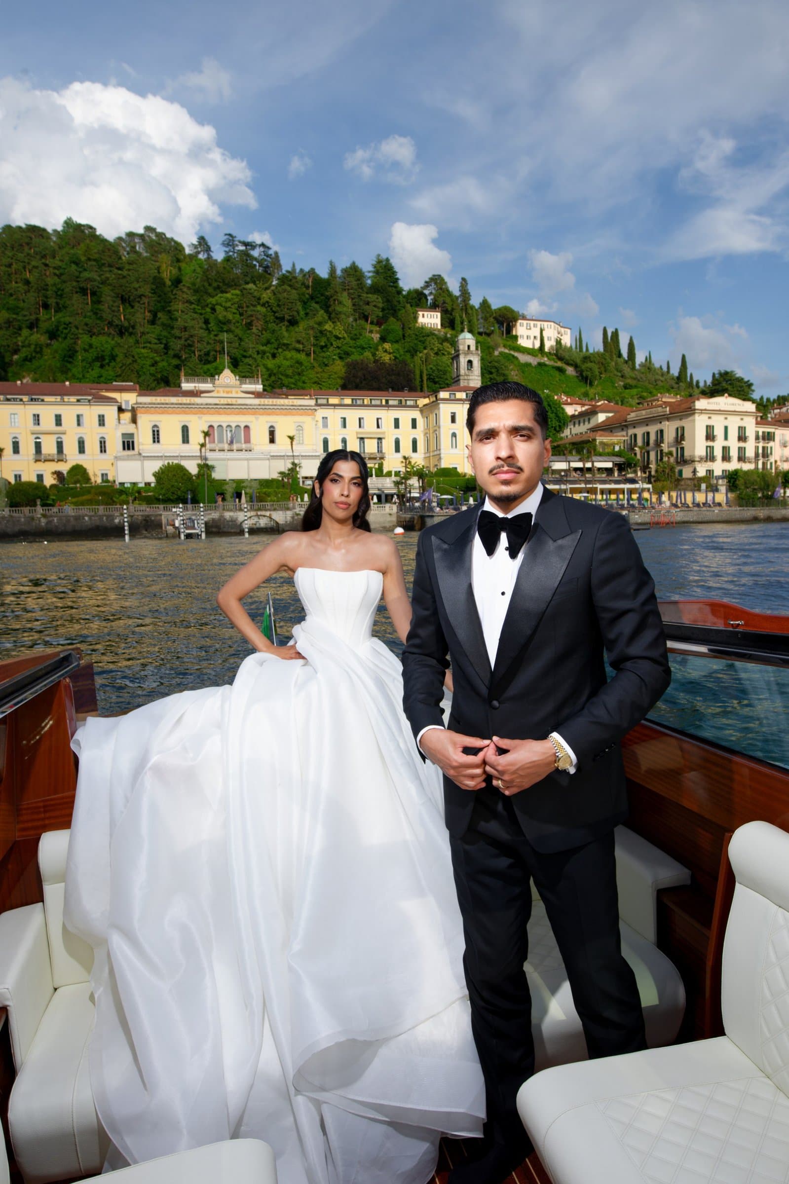 The groom in a black tuxedo beside the bride aboard a boat, a lakeside town behind