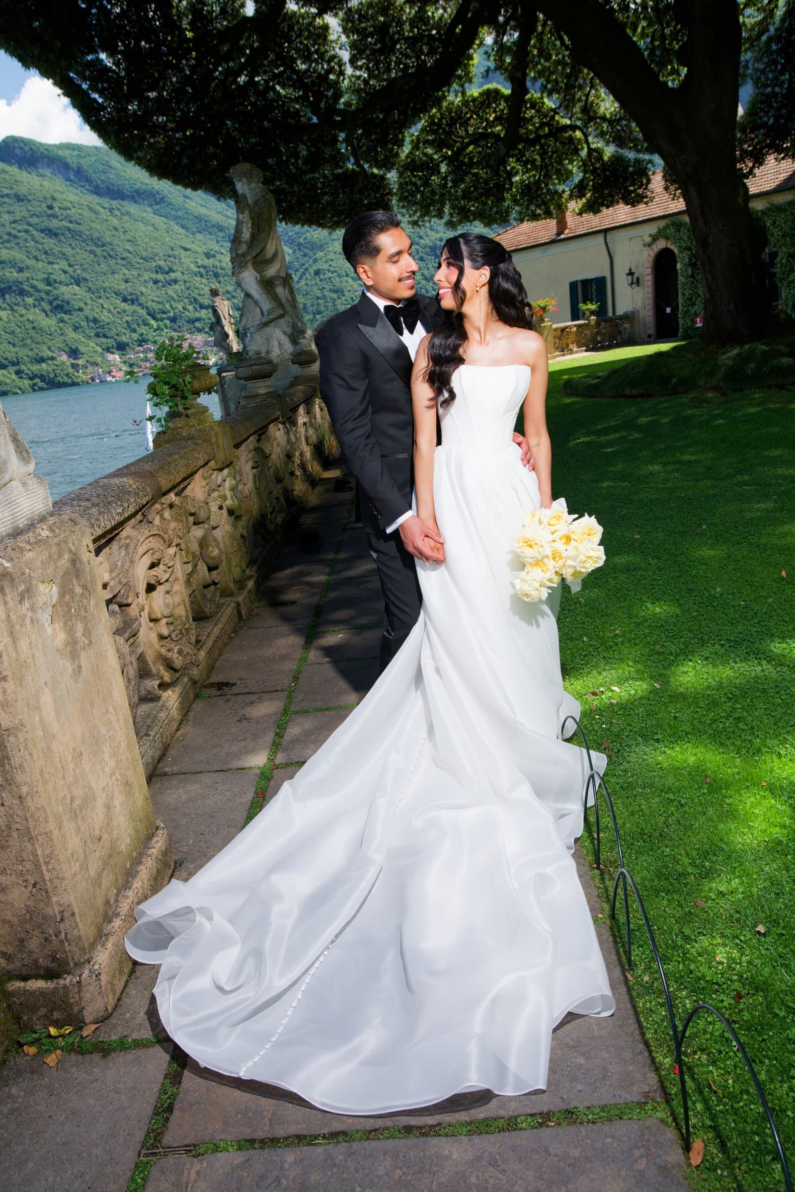 The bride and groom on a stone garden walk beside the villa lawn, she holding white roses
