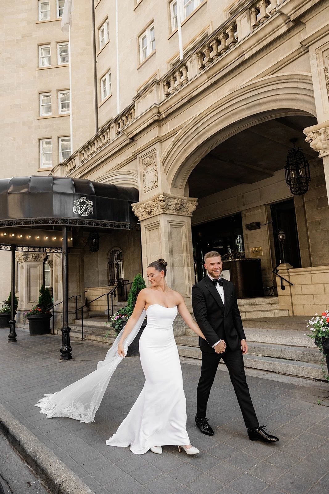 Couple at a stone hotel entrance, the groom in a black wedding suit, tailored in Edmonton